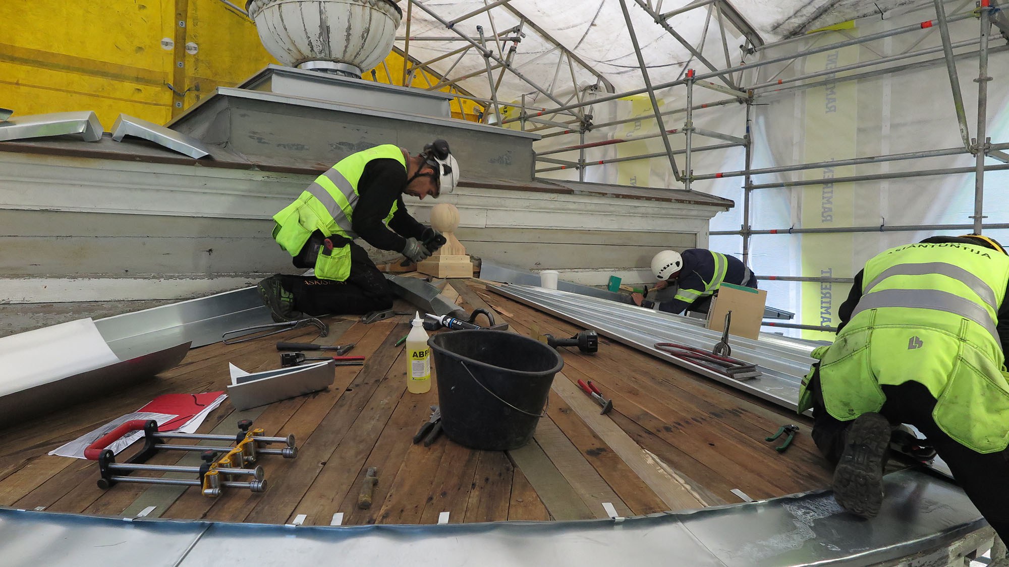 Puolipyöreän katon päällä työntekijöitä töissä pukeutuneena neonkeltaiseen liiviin ja turvakypärään.