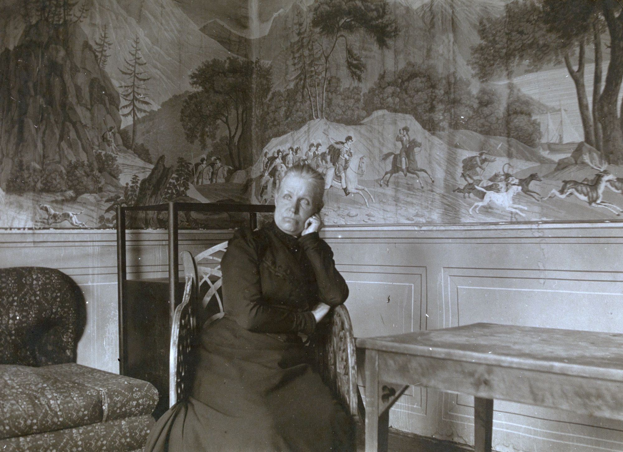 Mustavalkokuvassa Jenny Florin istuu tuolilla huvimajassa. Hänen takanaan näkyy kalusteita sekä huvimajan seinän panoraamatapettia.