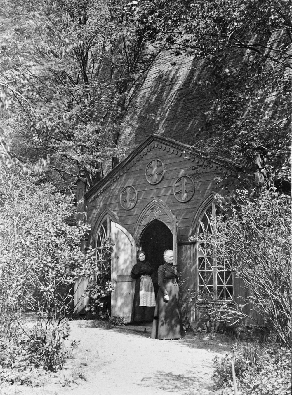 Mustavalkokuvassa kaksi naista seisoo huvimajan edustalla. Huvimajan ulko-ovet ovat auki. Etualalla on puita.